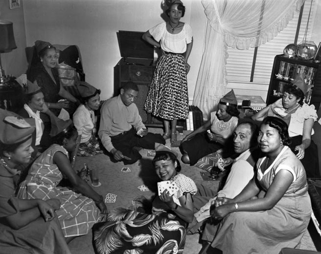 Women, children and two men seated around a living room playing cards. The women wear identical party hats (might this be a birthday?). Identifications: Possibly Glenn Buxton, Cecilia "Babe" Homes, Margie Henderson, Bruce Rowels, Madelaine Brown. CREDIT MOHAI, AL SMITH COLLECTION, 2014.49 