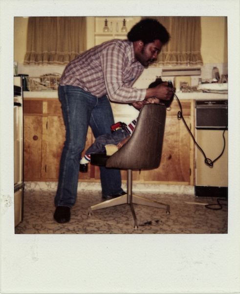 A father trims his son's afro hair style in the kitchen. Date Unknown.